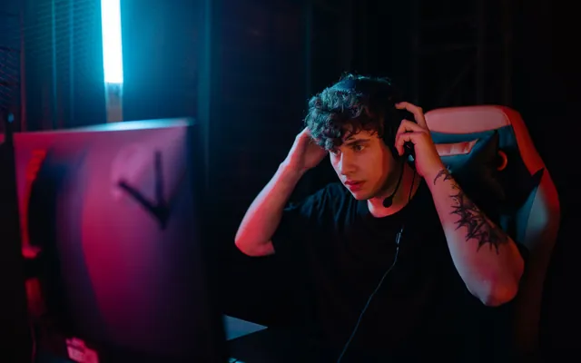 A streamer wearing a headset at a dark desk setup, lit by monitor glow.